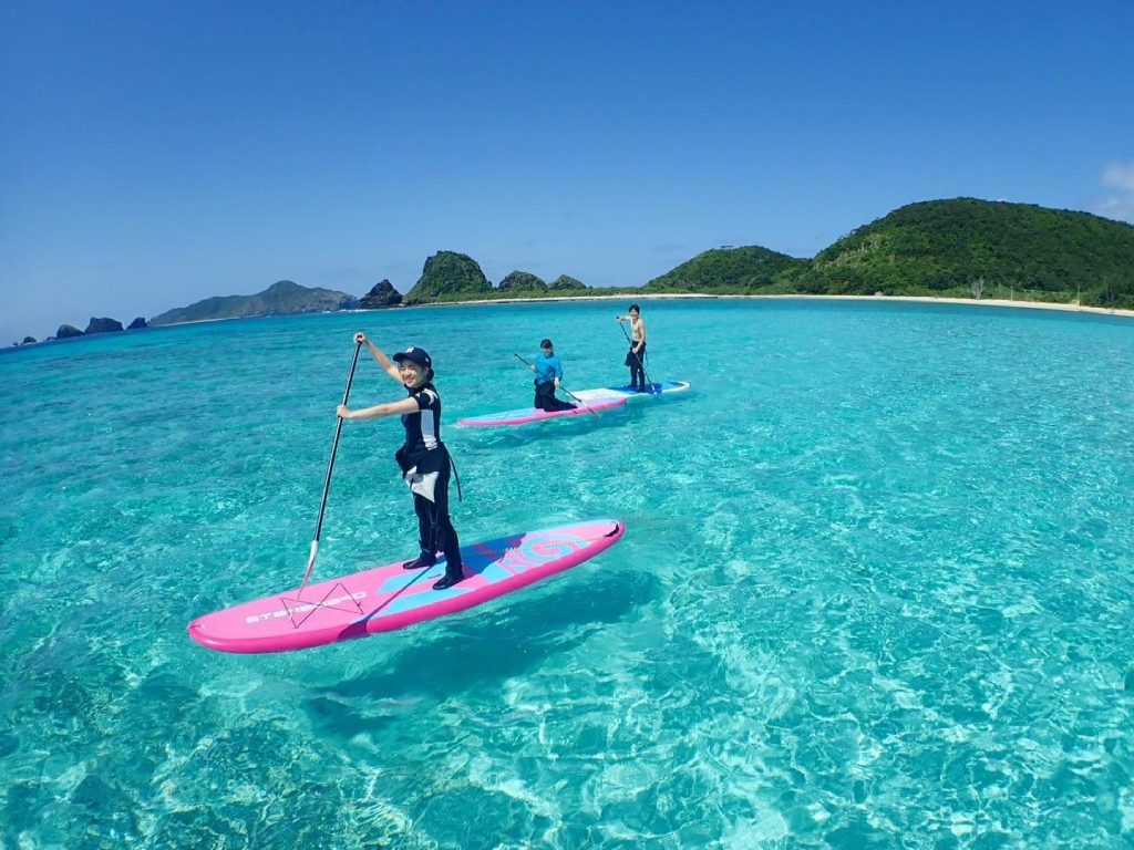 沖縄の透明な海の上で、3人がスタンドアップパドルボードに乗り、青い海と緑の島を背景に楽しんでいる様子
