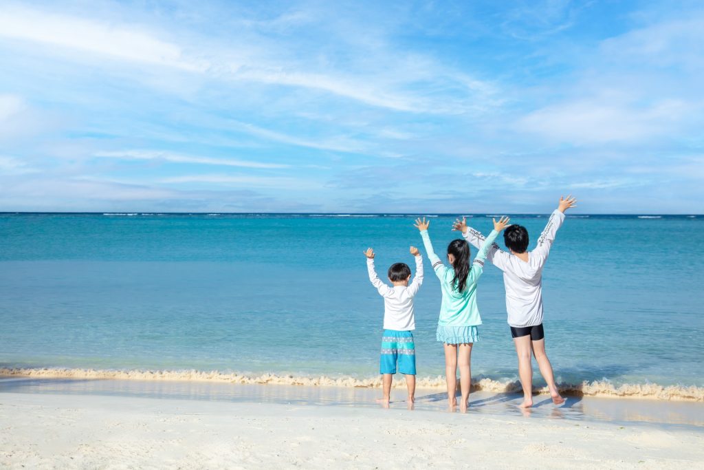沖縄の青い海と白い砂浜を前に、子ども2人と大人1人が並んで立ち、両手を上げて景色を楽しんでいる家族の後ろ姿